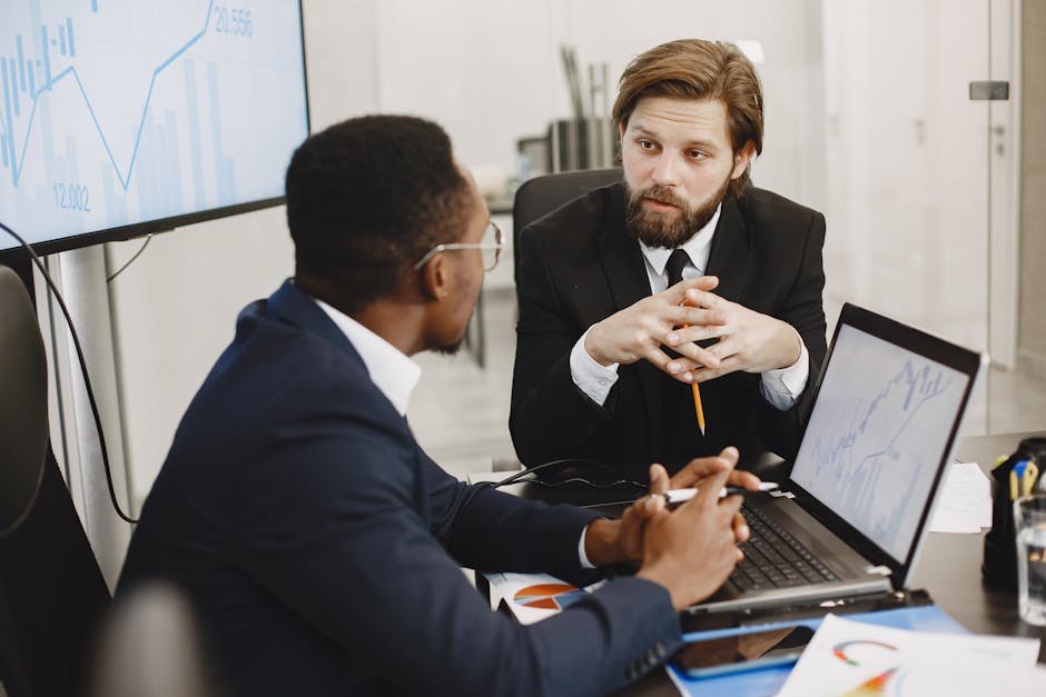 Two businessmen in suits discussing financial data with laptops in a modern office setting.